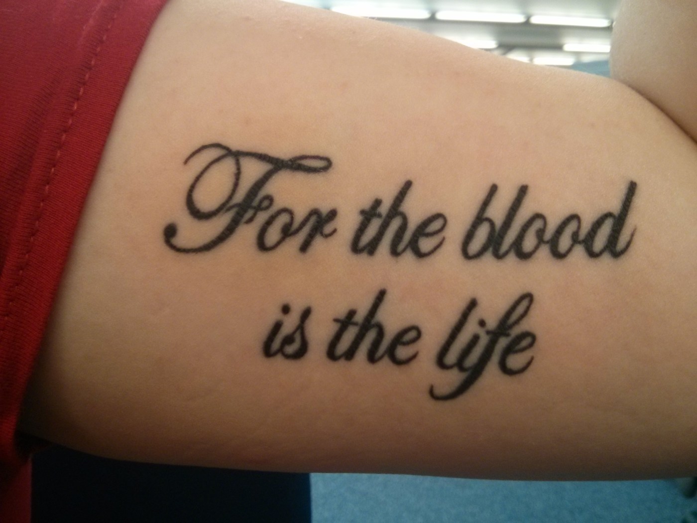 Image shows a written tattoo on a bicep. It reads "For the blood is the life" in curling script.