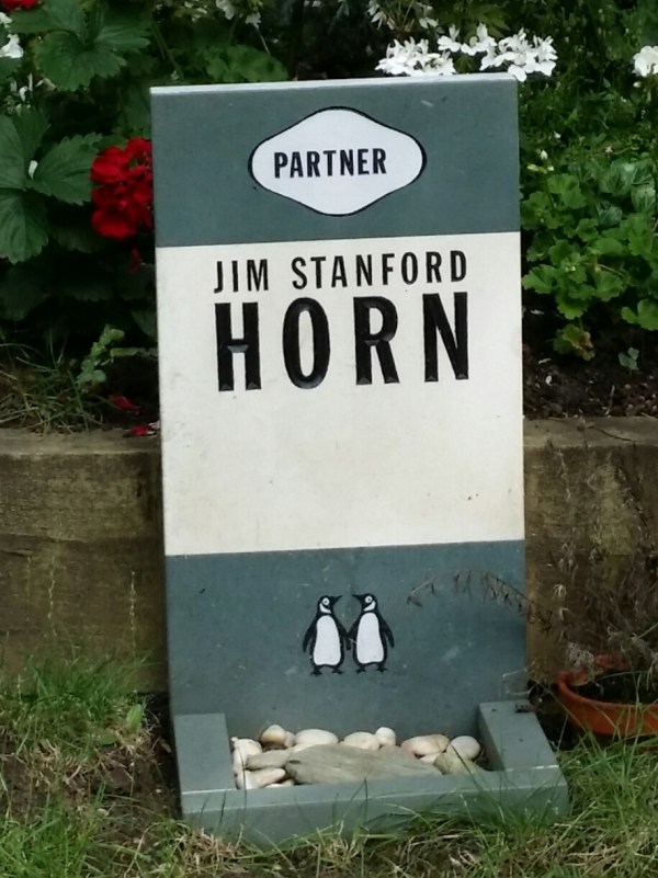 The image shows a gravestone designed to look like a Penguin book cover. It has a blue stripe at the top and bottom and white one in the middle. At the top is the word "Partner". In the middle is the name "Jim Stanford Horn". As the bottom are two penguins holding wings.
