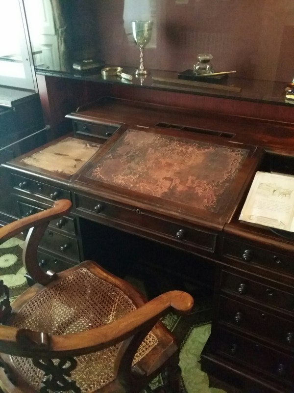 The image shows Charles Dickens writing desk. It is a wooden desk with drawers and a mottled writing pad in the middle. In front of it is a wooden chair with a reed seat.