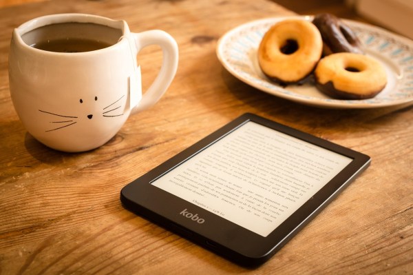 A mug with a cat's face sits on a table next to an e-reader and plate with doughnuts