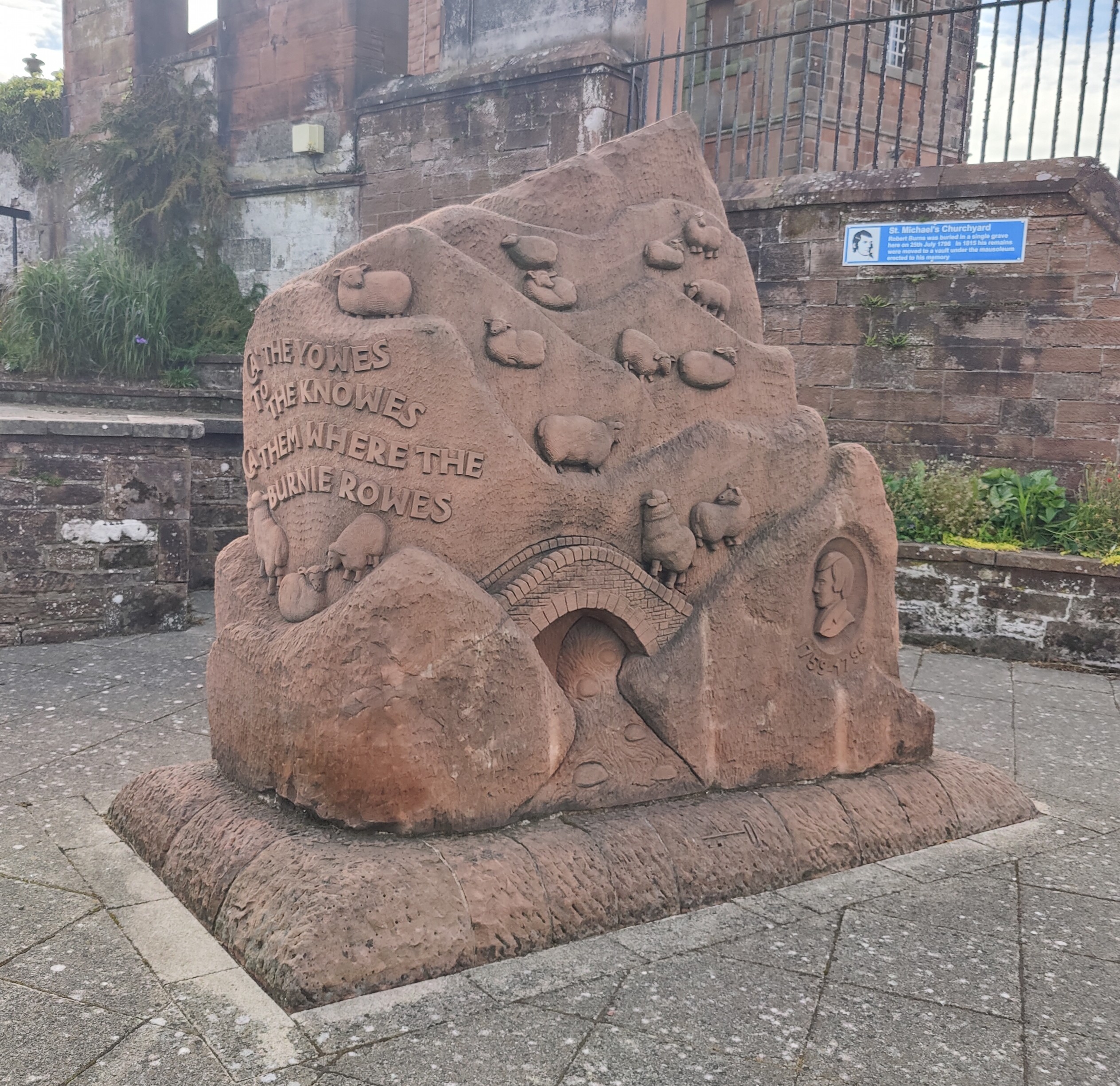 A large red sculpture stands in front of a brock wall. It is shaped to look like a hill. On it, various sheep walk on the hill, one crossing a bridge. Writing reads "Ca the Yowes to the knowes, ca them where the burnie rowes"